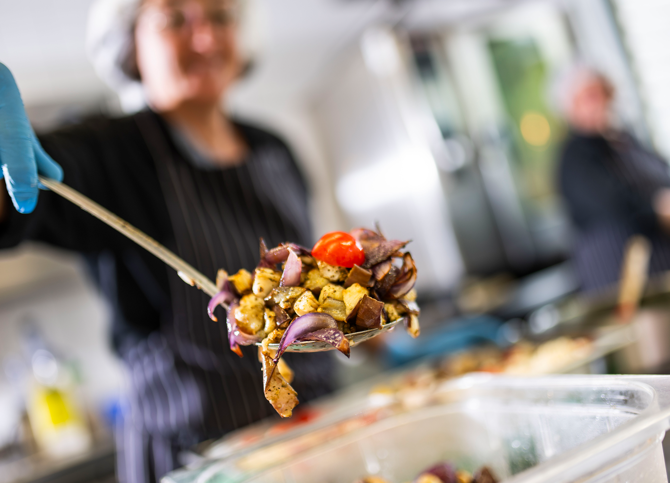 Küchenmitarbeiterin hält eine Kelle mit gebratenem Gemüse über einer Buffetbox